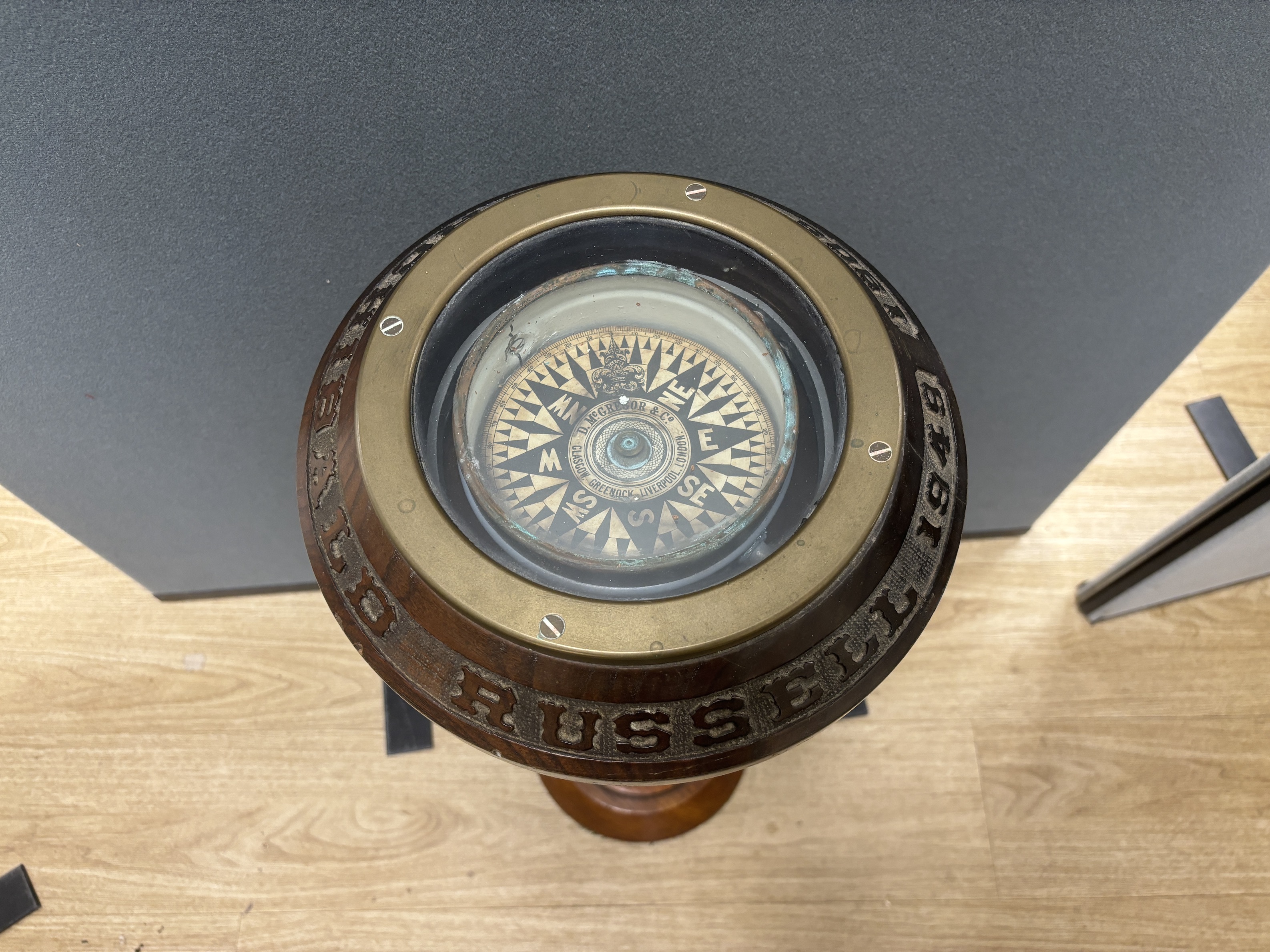 A ship’s compass in a gimbal, believed to be from the tall ship The Archibald Russell, mounted on a floor standing mahogany stand with carved attribution around the edge, together with a ship in a bottle of the barque in